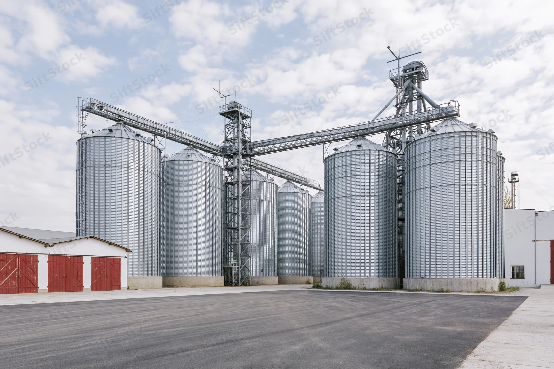 Large grain elevator complex with multiple tall corrugated metal silos and conveyor system under partly cloudy sky