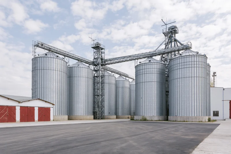 Large grain elevator complex with multiple tall corrugated metal silos and conveyor system under partly cloudy sky