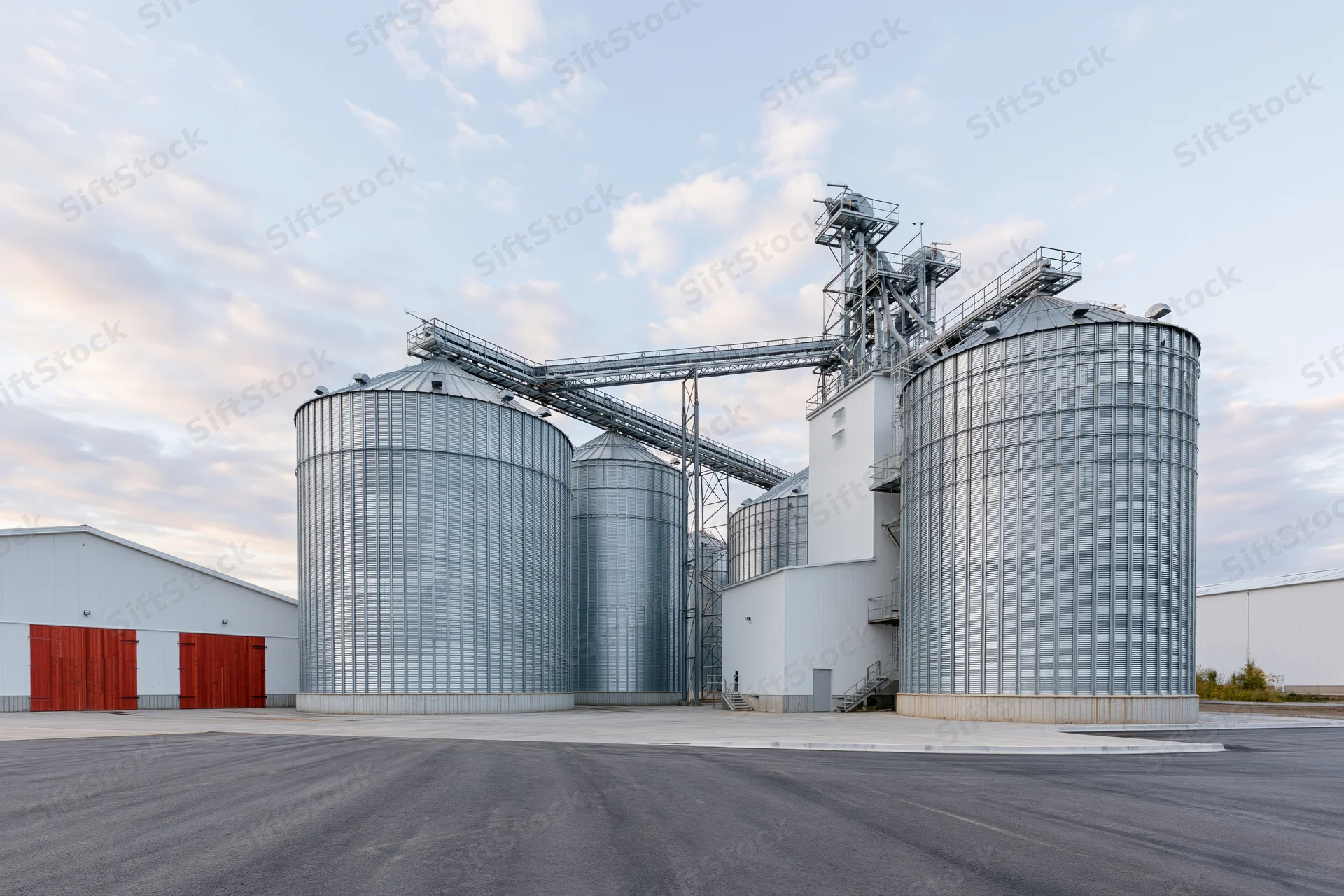 Large grain elevator complex with multiple tall corrugated metal silos and conveyor system under daytime sky