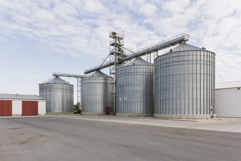 Large grain elevator complex with multiple tall corrugated metal silos under a partly cloudy sky