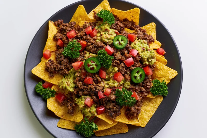 Loaded nachos with beef, guacamole, tomatoes, jalapenos and parsley on a dark round plate viewed from above
