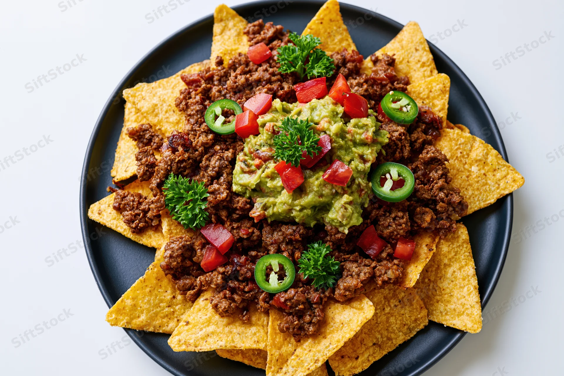 Loaded nachos with beef, guacamole, tomatoes, jalapenos and parsley on a dark round plate viewed from above