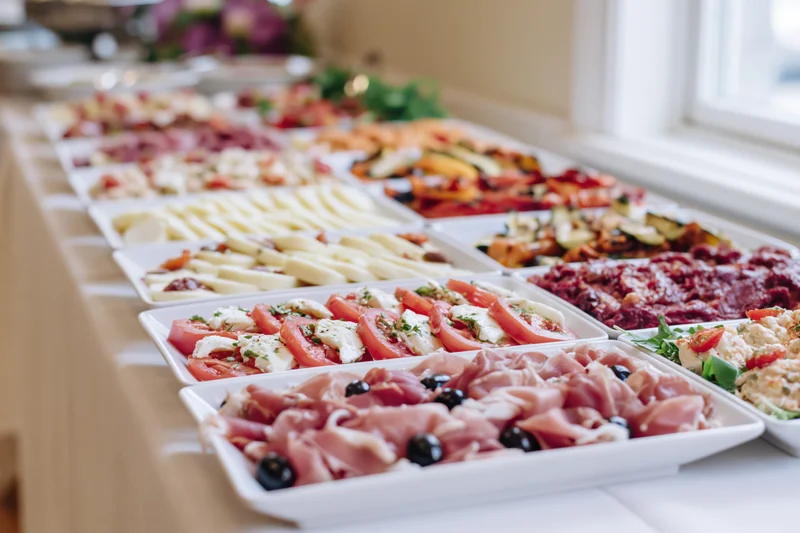 Long table with white linen holding multiple platters of cold appetizers and salads arranged in rows near a window