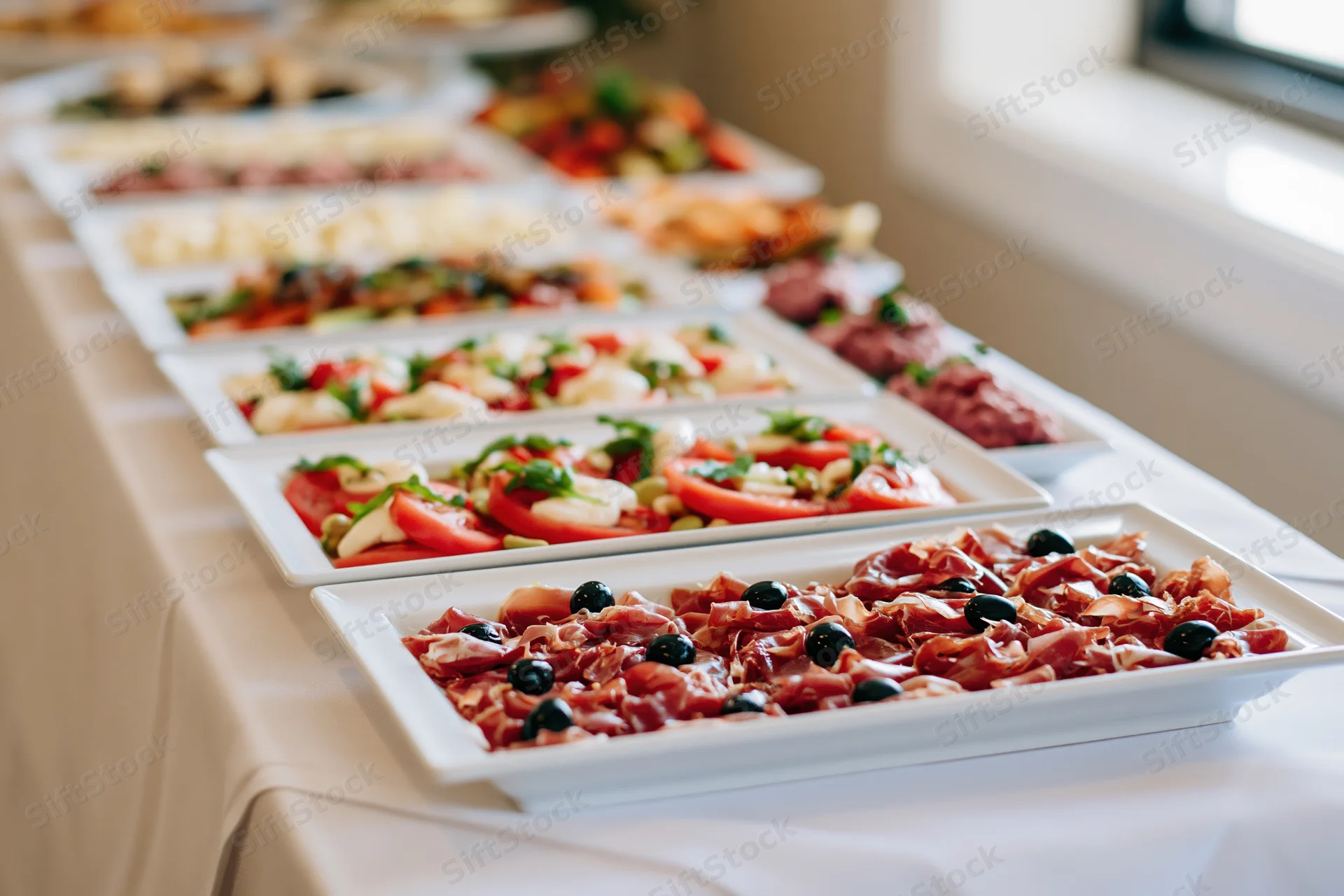 Long banquet table with white linen holding multiple platters of appetizers and salads arranged in rows for a reception event