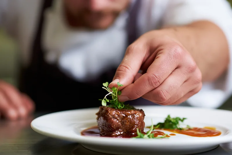 Chef placing microgreen garnish on plated steak in restaurant kitchen