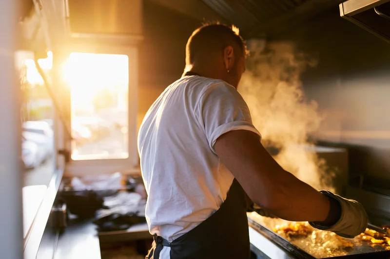 Male chef cooking on a flat-top griddle in a compact kitchen with steam rising and sunlight streaming through a window