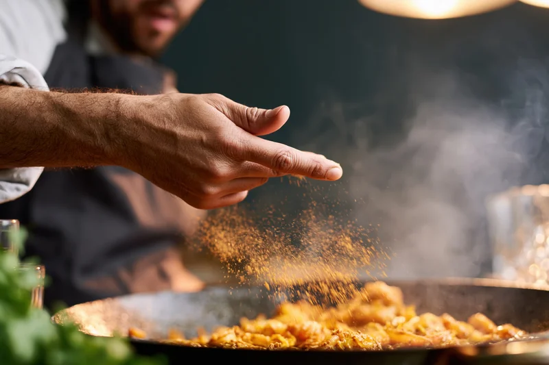 Chef sprinkling spice into steaming dish in kitchen