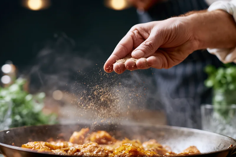 Chef's hand sprinkling ground spice into steaming pan of frying food in kitchen