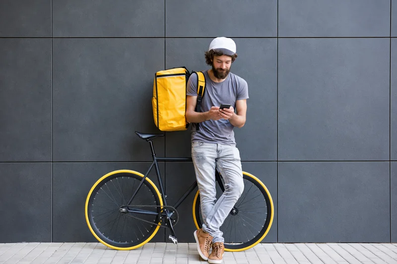 Delivery courier leaning against tiled wall with bicycle, checking smartphone