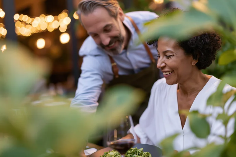 Waiter serving a woman at an outdoor restaurant table with foliage framing the scene