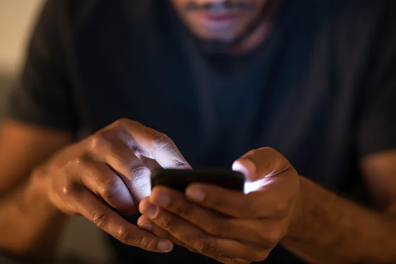 Man tapping smartphone screen with both hands in dim indoor setting