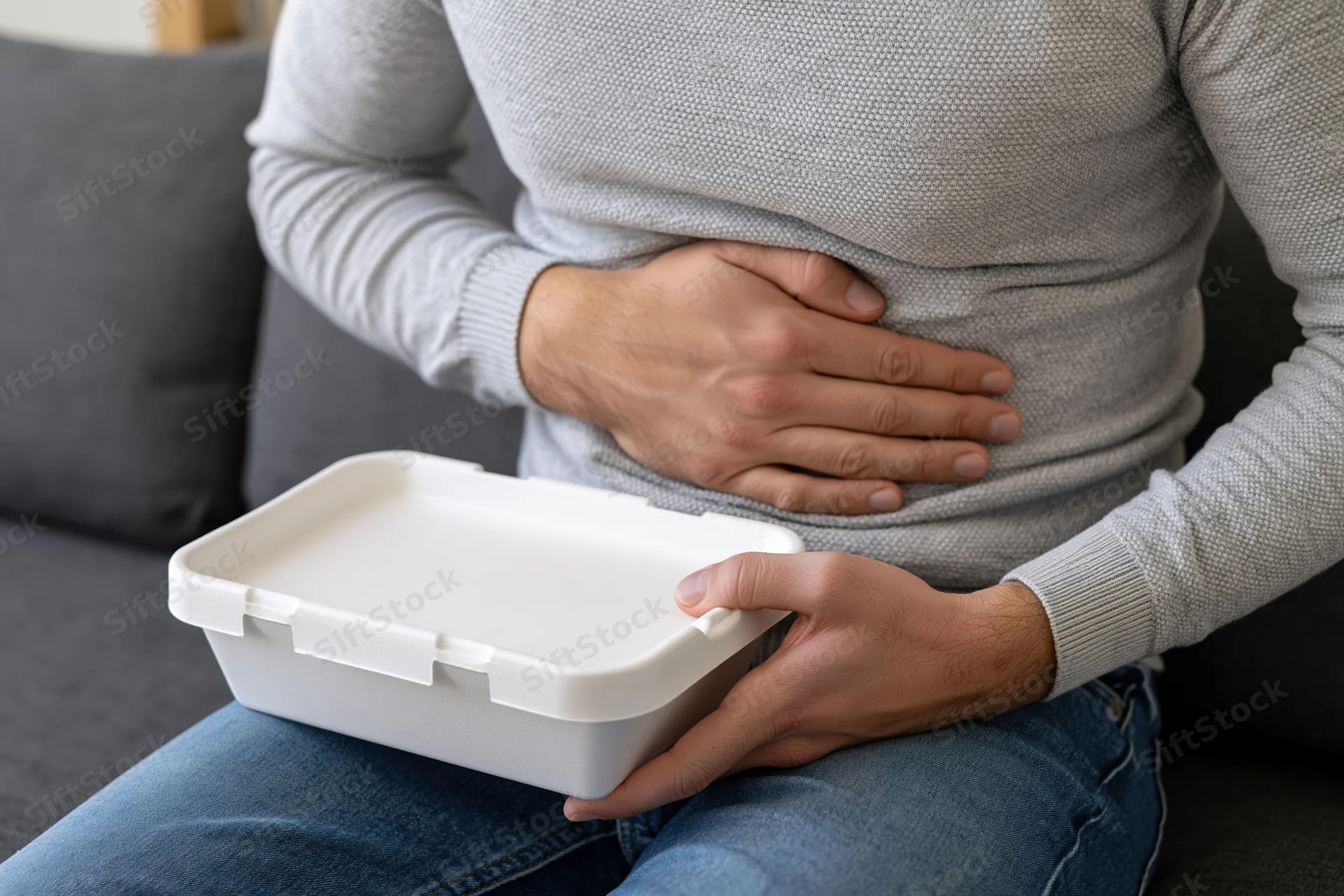 Man holding styrofoam food container while clutching his stomach in discomfort on a couch