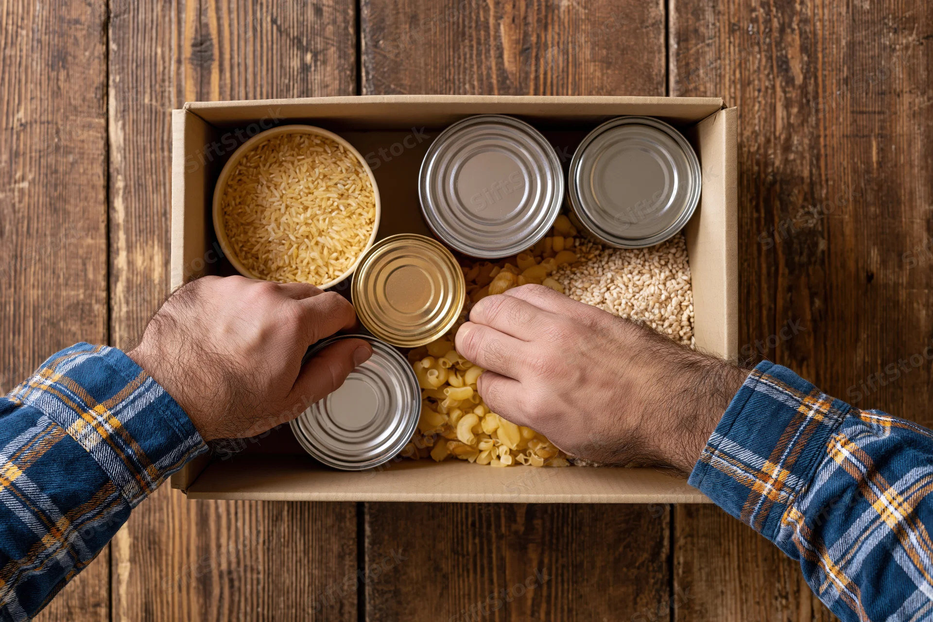 Man arranging canned goods and dry staples in a cardboard box on wooden surface
