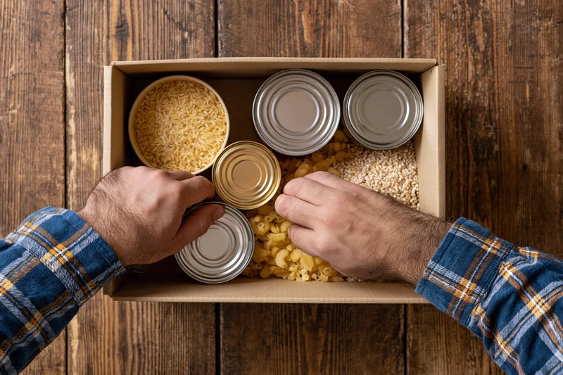 Man arranging canned goods and dry staples in a cardboard box on wooden surface