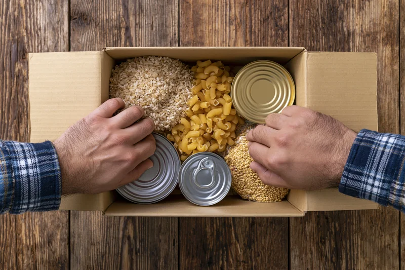 Man’s hands arranging canned goods and dry staples in a cardboard box on wooden surface