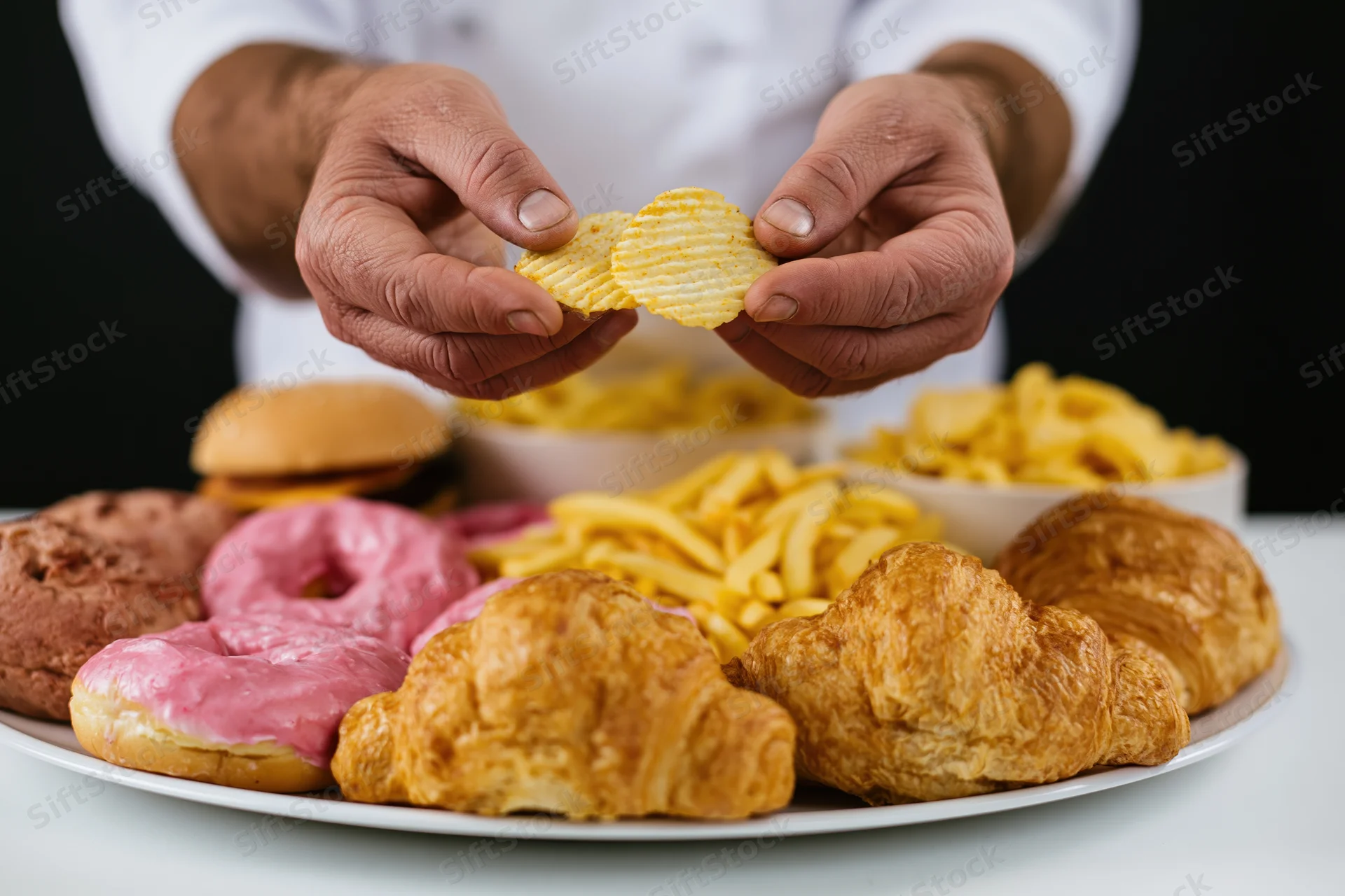 Hands holding ridged potato chips above a plate of assorted snacks on a white table