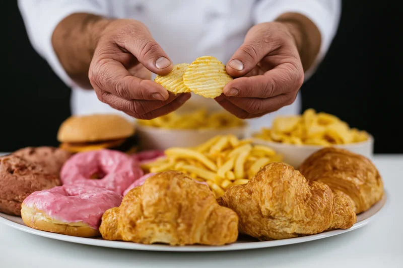 Hands holding ridged potato chips above a plate of assorted snacks on a white table