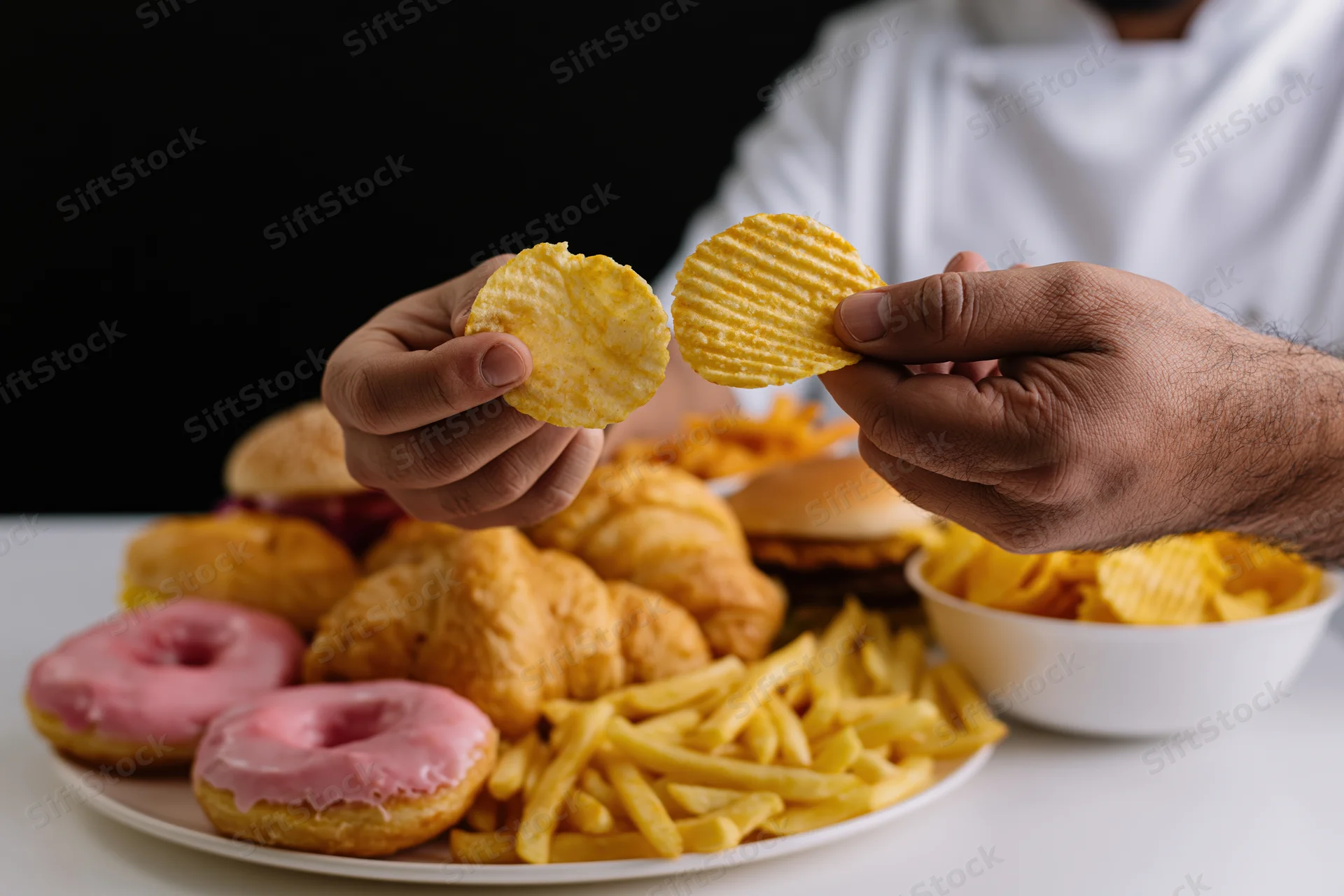 Man holding two ridged potato chips above a table with donuts, fries, croissants and burgers