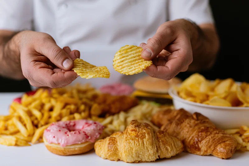 Man holding two ridged potato chips above a table with fries, donut, croissants and chips