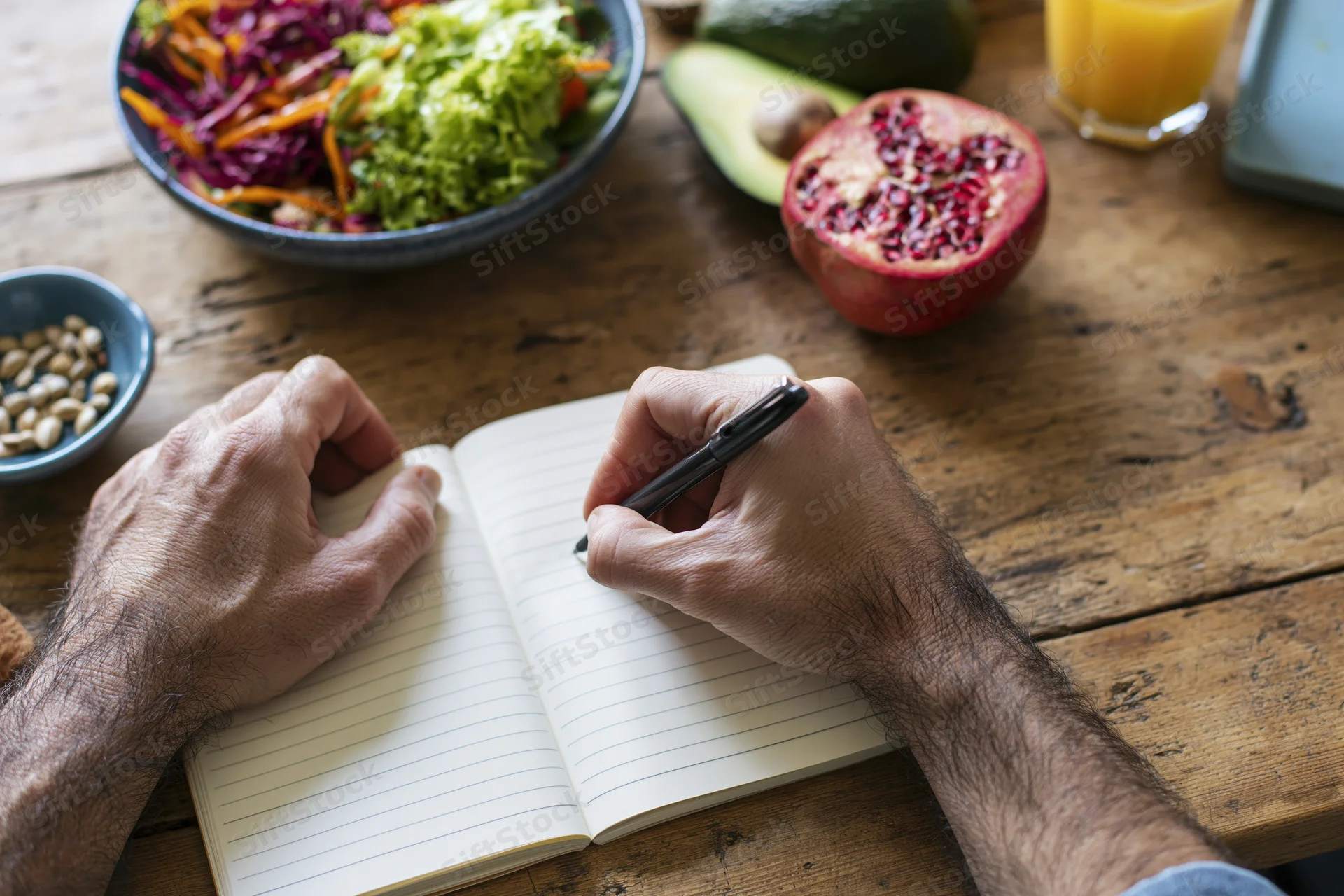 Man's hands writing in a lined notebook on a rustic wooden table with fresh salad and fruit nearby