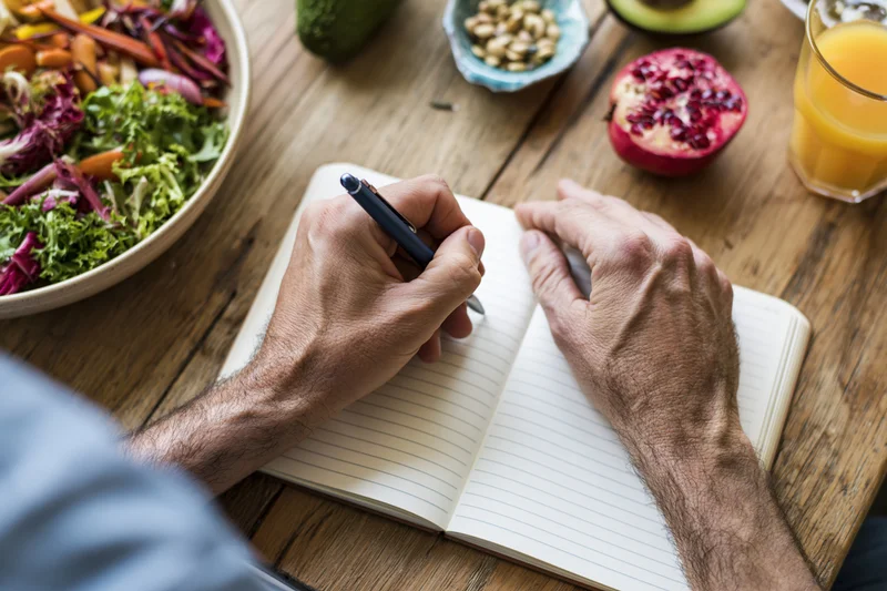 Man's hands writing in a lined notebook on a rustic wooden table with fresh food nearby