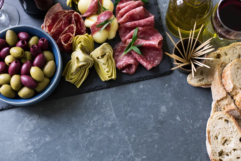 Mediterranean antipasto spread with cured meats, olives, artichokes, bread and wine on a dark slate surface