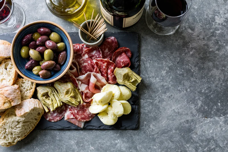 Mediterranean antipasto spread with cured meats, cheese, olives and bread on a dark slate board with wine glasses and olive oil bottle nearby