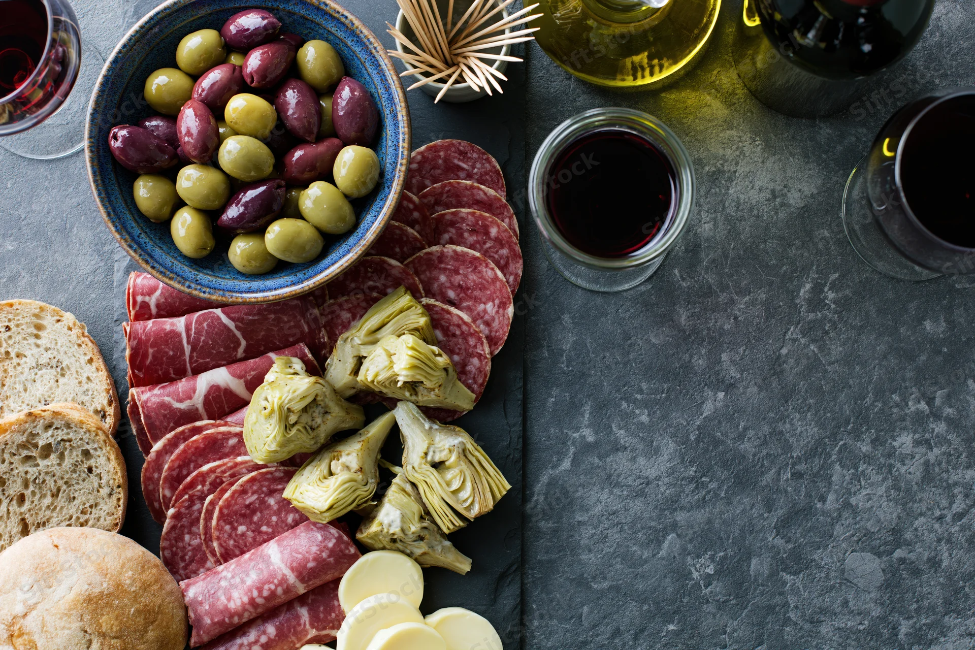 Antipasto spread with salami, olives, artichokes, cheese and bread on a dark slate surface