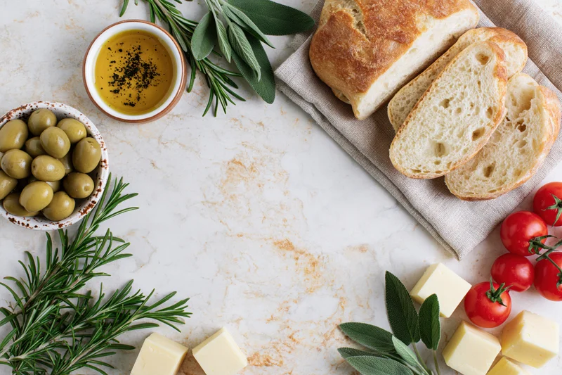 Mediterranean food flat lay arranged in loose border around marble surface with bread, olives, tomatoes, cheese, herbs and olive oil