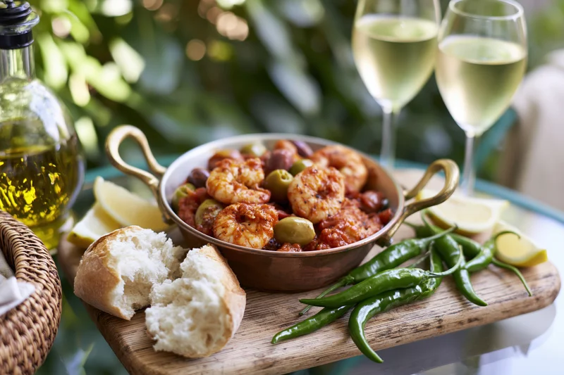 Shrimp in tomato sauce with olives and bread on a rustic wooden board outdoors with white wine and green peppers