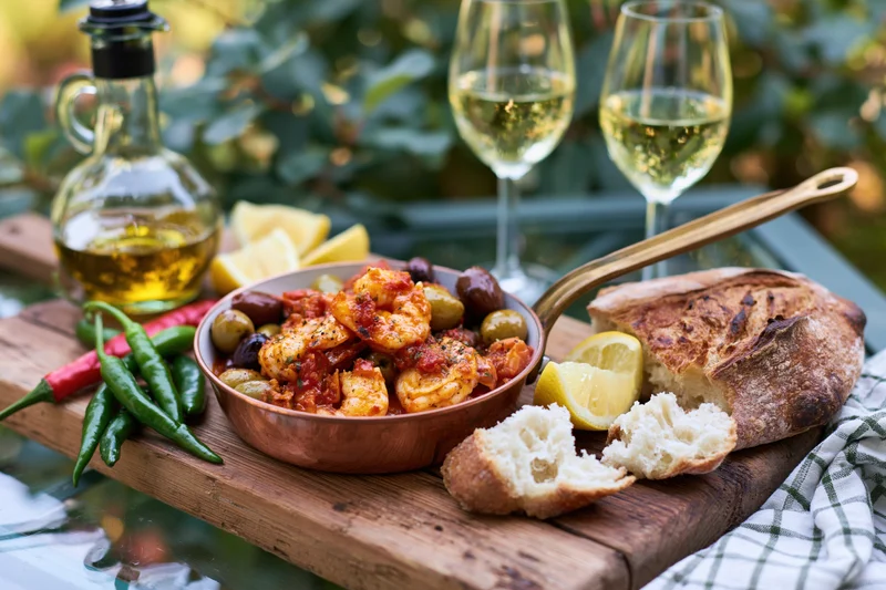 Shrimp and tomato dish with olives and bread on a wooden board outdoors with wine and lemon slices