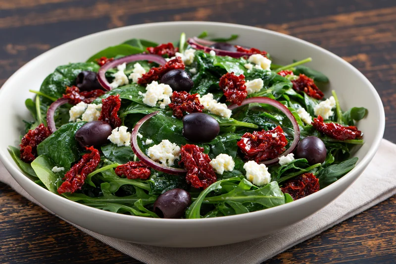 Mediterranean salad with arugula, spinach, kalamata olives, sun-dried tomatoes and feta in a white ceramic bowl on a dark wooden table
