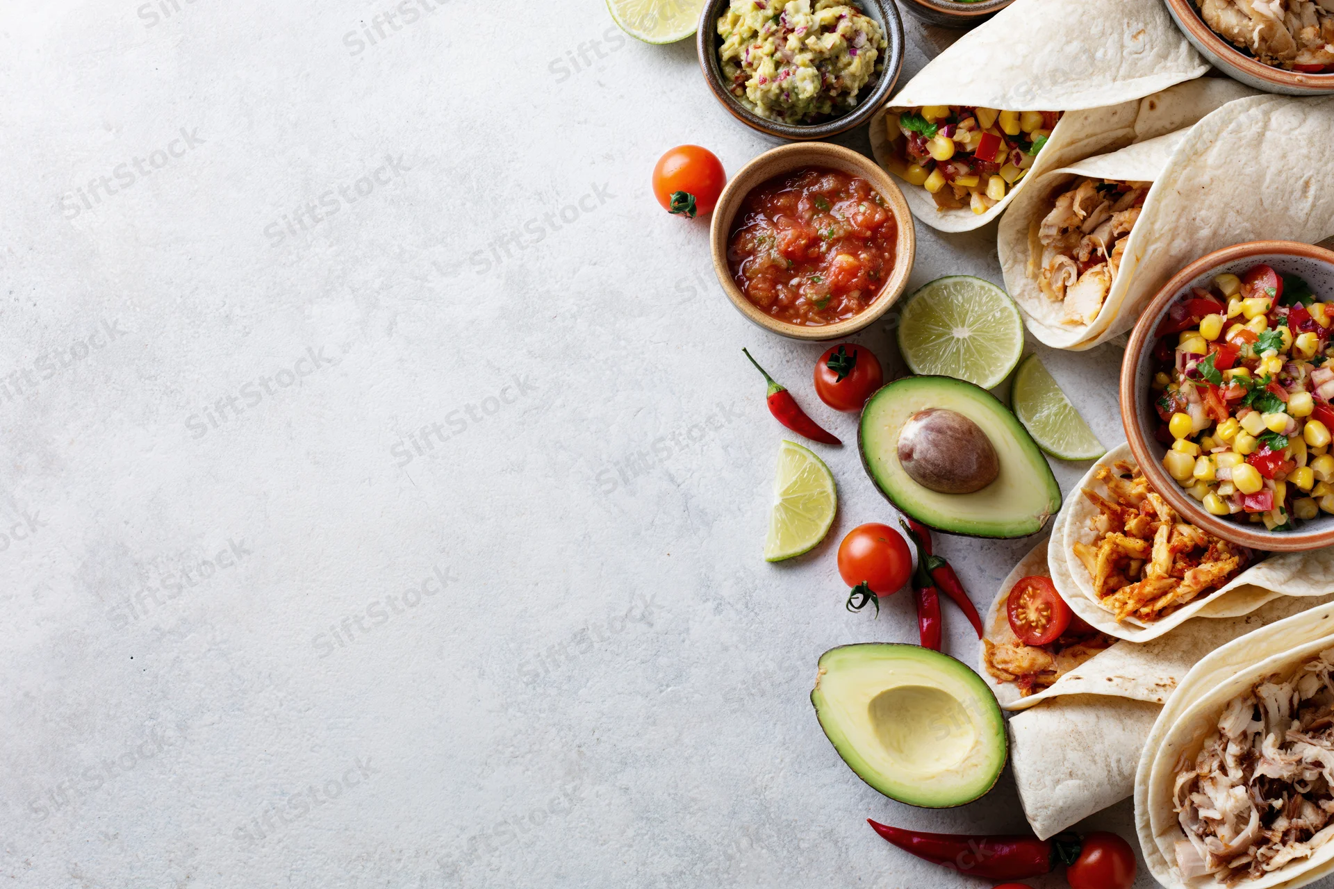 Mexican food spread with tacos, avocado, salsa and guacamole arranged along right edge of white surface