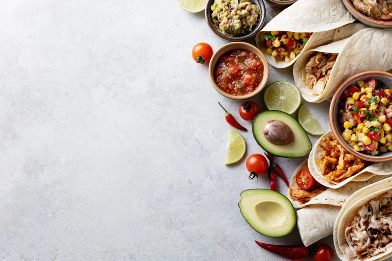 Mexican food spread with tacos, avocado, salsa and guacamole arranged along right edge of white surface