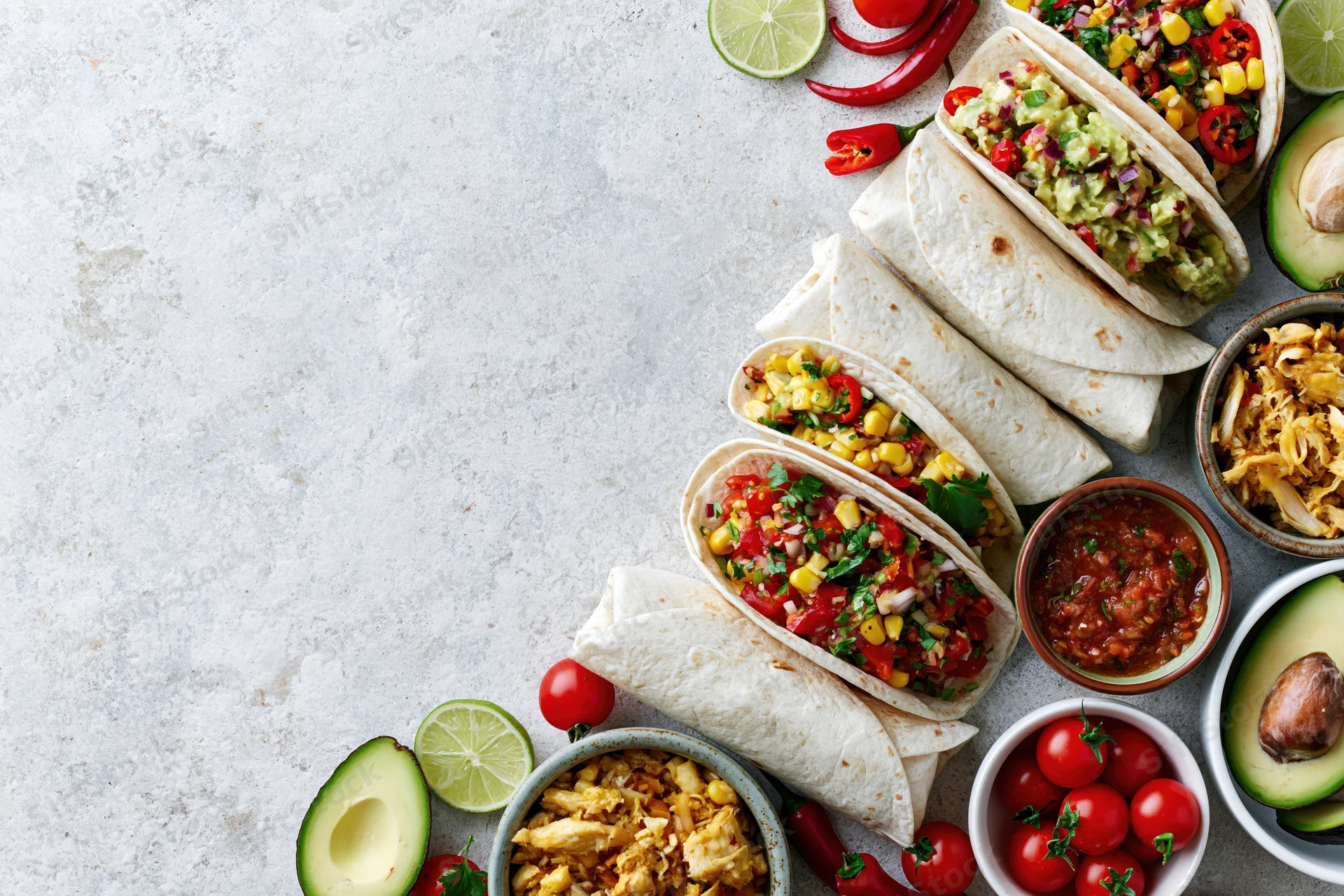 Mexican food spread with tacos, avocado, lime, and salsa arranged along right edge of white stone surface