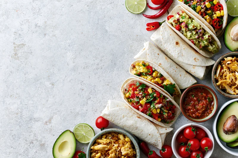 Mexican food spread with tacos, avocado, lime, and salsa arranged along right edge of white stone surface