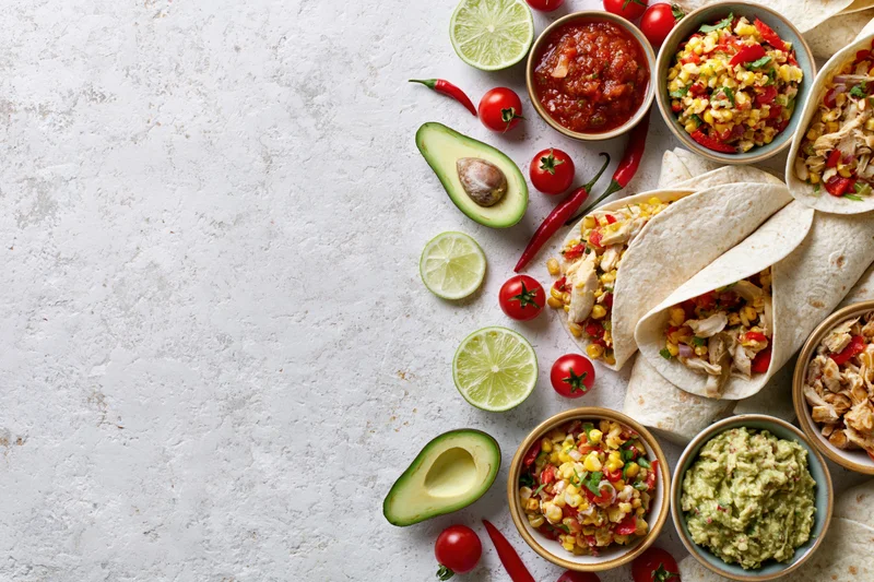 Mexican food spread with tacos, guacamole, salsa, and fresh ingredients arranged along the right edge of a white textured surface
