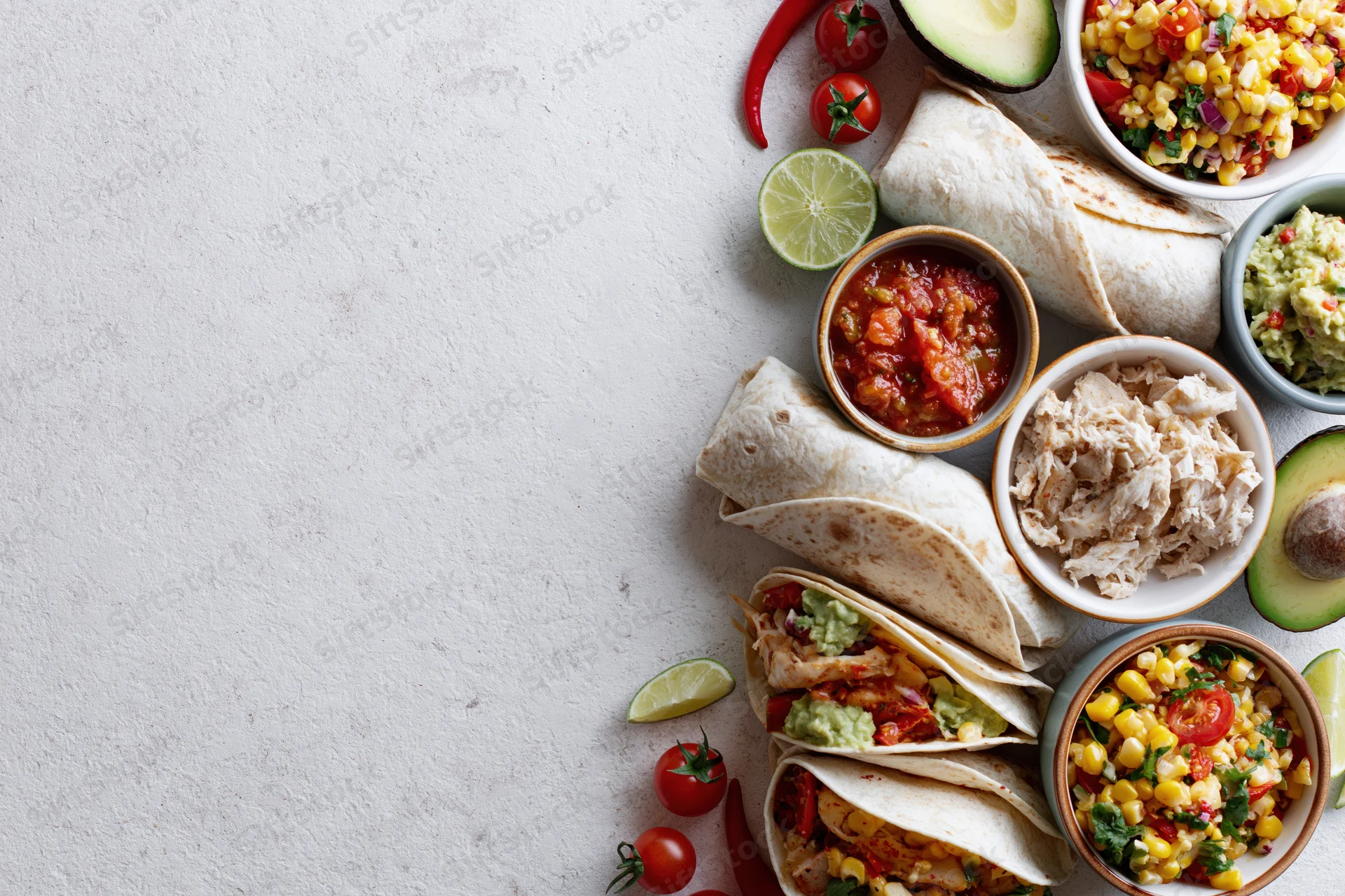 Mexican food spread with tacos, salsa, guacamole and fresh ingredients arranged along the right edge of a white surface