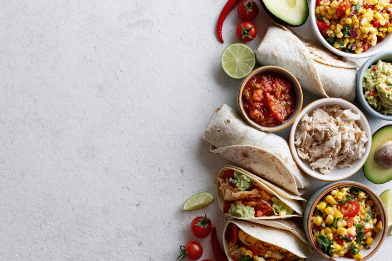 Mexican food spread with tacos, salsa, guacamole and fresh ingredients arranged along the right edge of a white surface