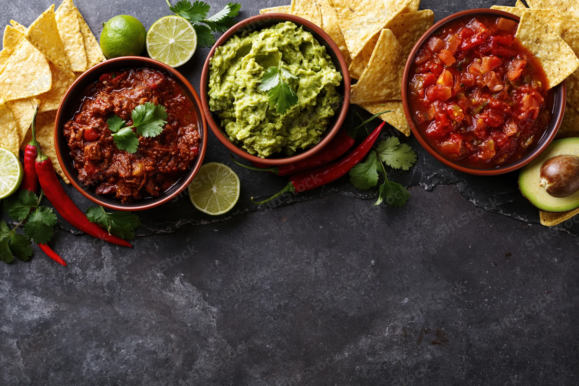 Mexican food spread with guacamole, salsa, chili, tortilla chips, lime, cilantro, and peppers arranged along the top edge of a dark stone surface
