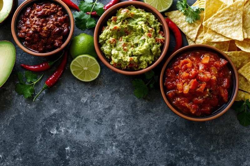 Mexican food spread with guacamole, salsa, refried beans, chips and lime arranged along top edge of dark stone surface
