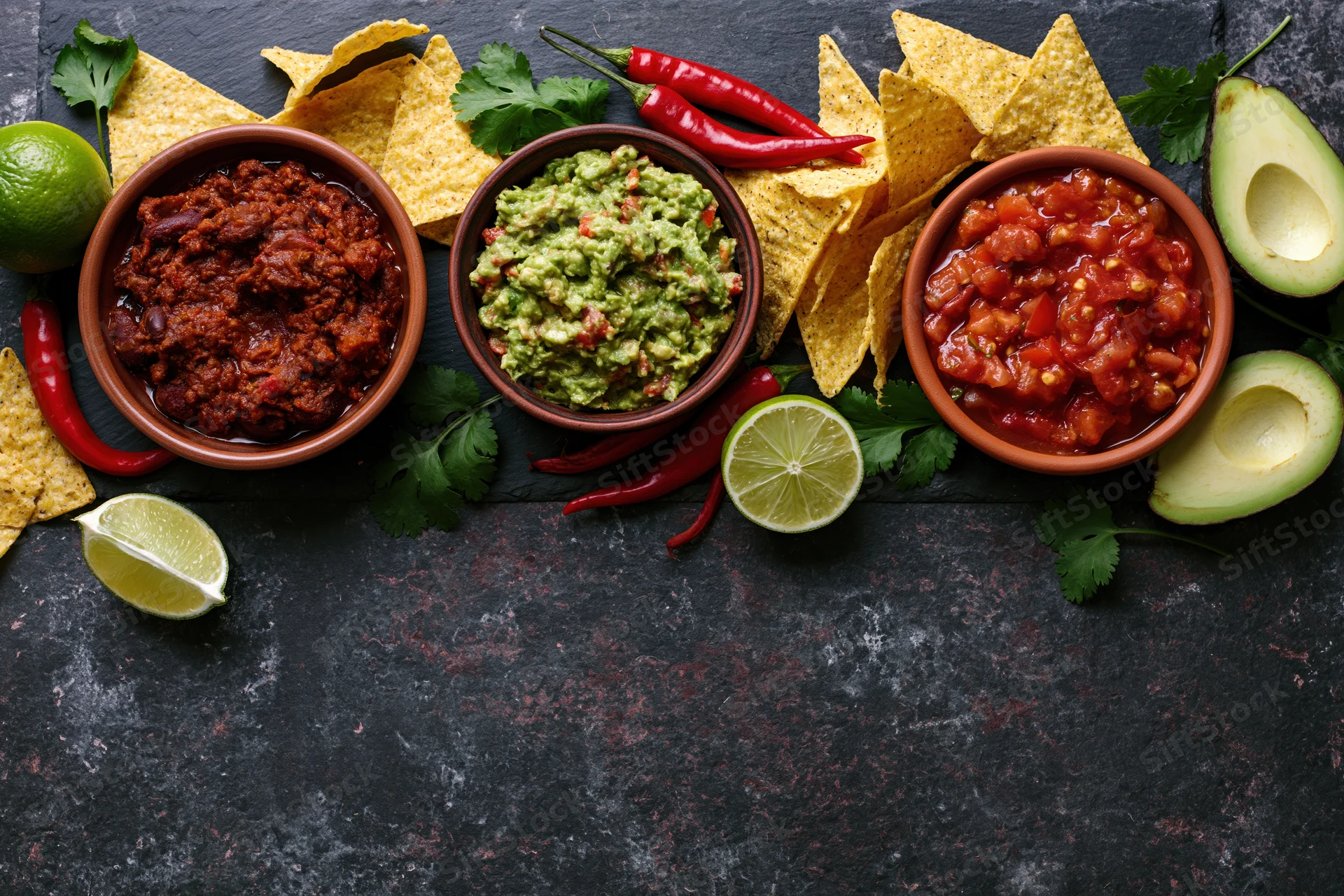 Mexican food spread with chili, salsa, guacamole, tortilla chips, avocado, lime and cilantro arranged along dark surface with copyspace below