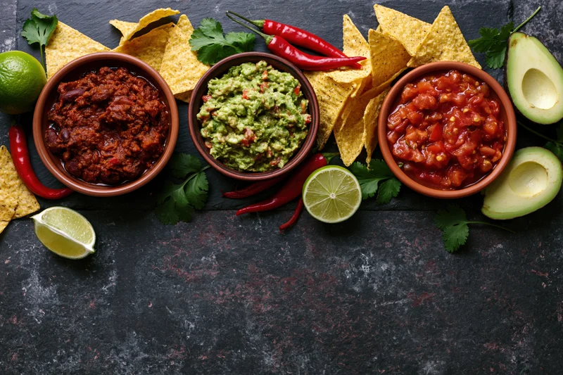 Mexican food spread with chili, salsa, guacamole, tortilla chips, avocado, lime and cilantro arranged along dark surface with copyspace below