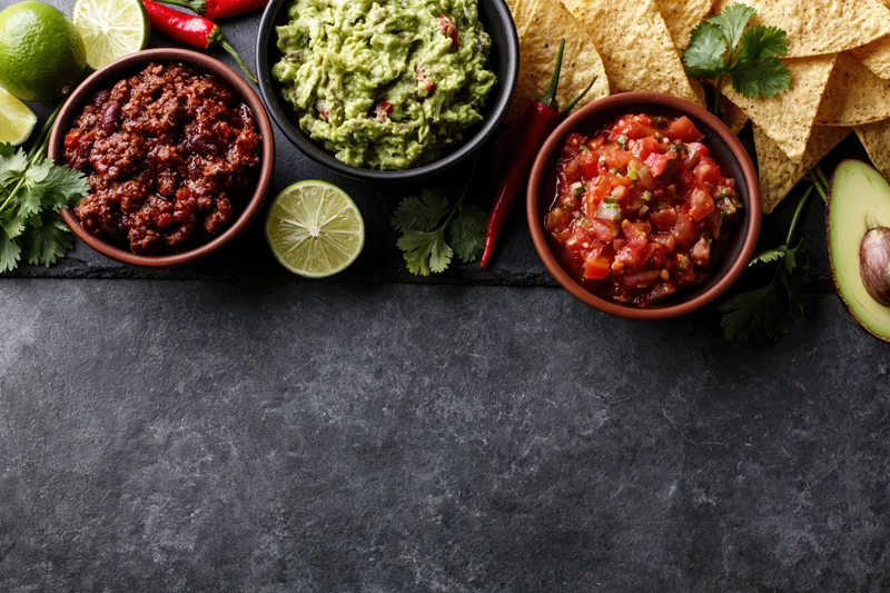 Mexican food spread with guacamole, salsa, refried beans, tortilla chips, lime, cilantro and chili arranged along top edge of dark slate surface
