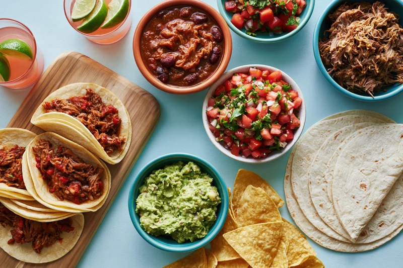Overhead view of a mexican food spread with tacos, guacamole, salsa, beans, chips and lime drinks on a blue surface