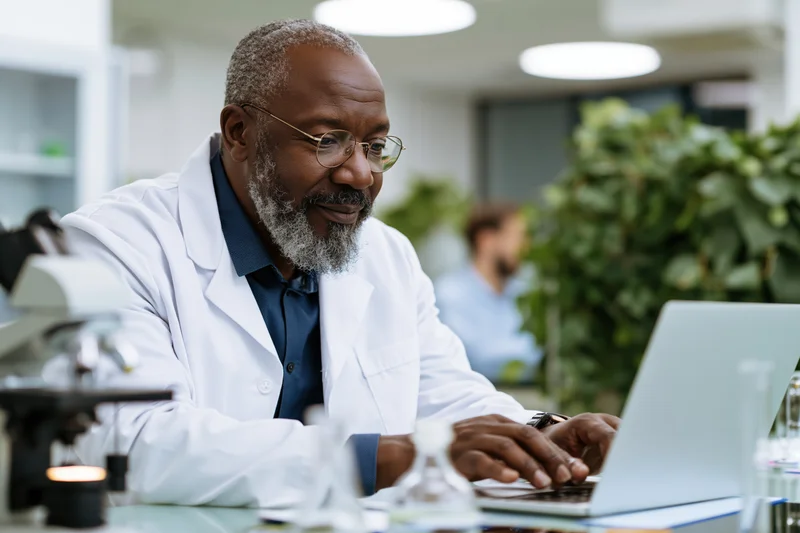 Middle-aged black male scientist in white lab coat typing on laptop in laboratory workspace