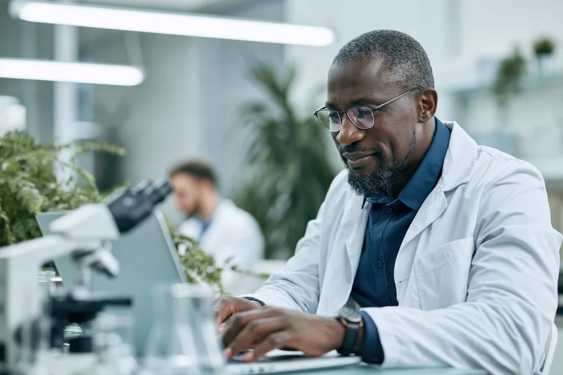 Middle-aged black male scientist in white lab coat typing on laptop at lab bench with microscope and glassware nearby