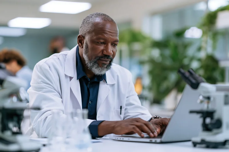 Middle-aged black male scientist in white lab coat typing on laptop at lab desk
