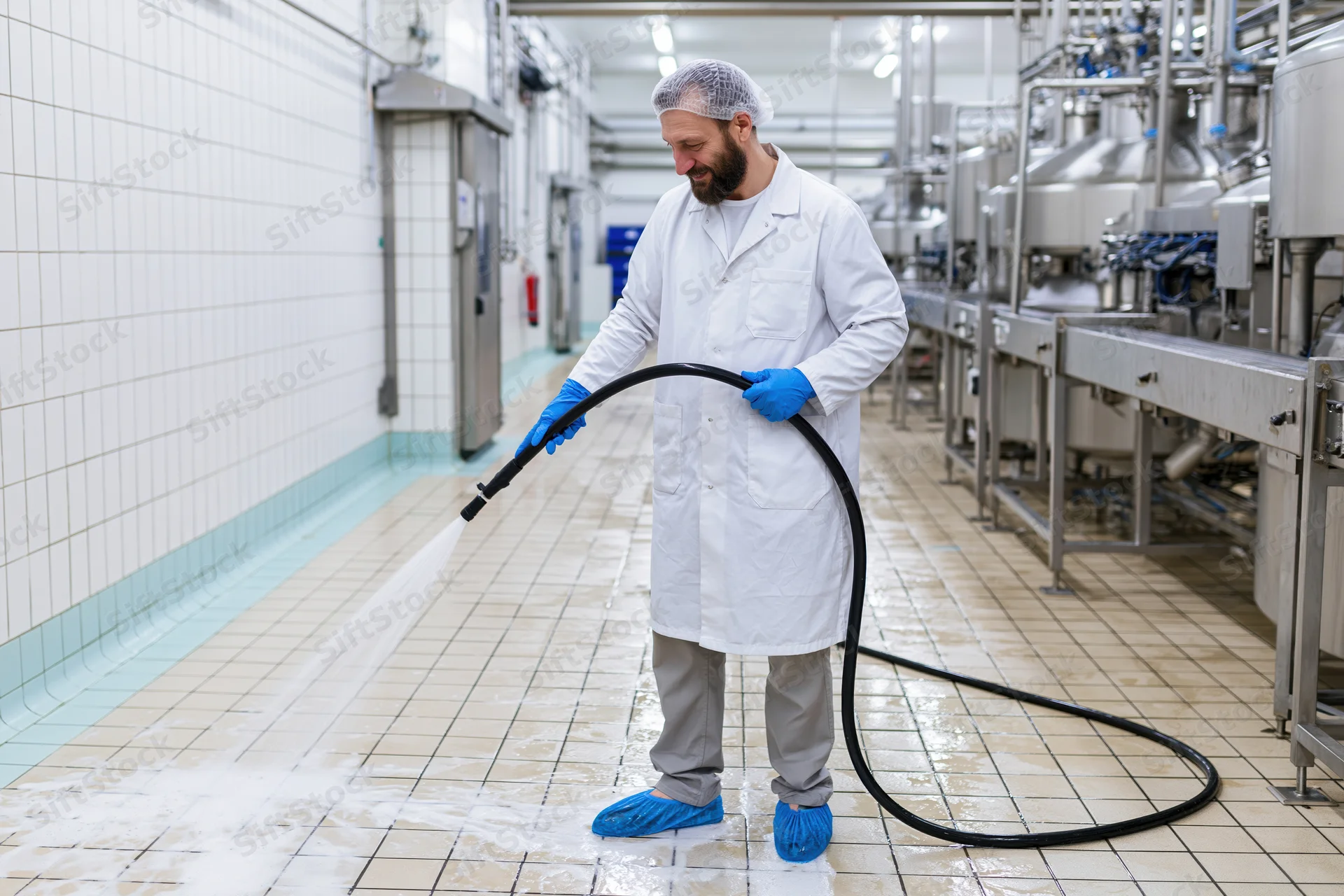 Man in lab coat and hairnet hosing down tiled floor in industrial food processing facility