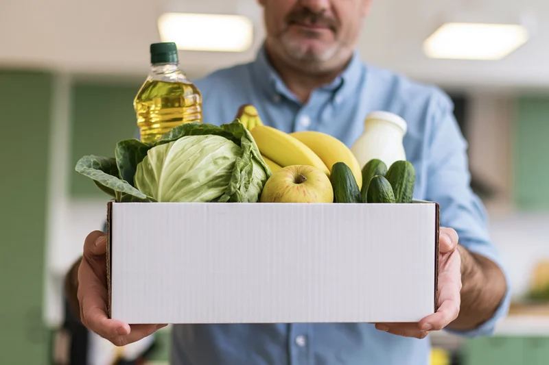 Man holding a white cardboard box filled with fresh produce and groceries in a kitchen